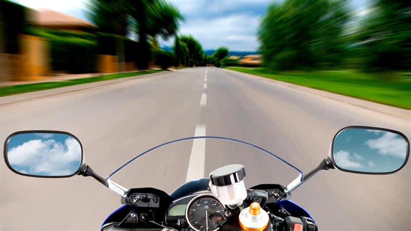 Kuvan kohteena moottoripyörän ohjaustanko ja mittaristo. Taustalla näkyy sumeasti tie ja maisema.