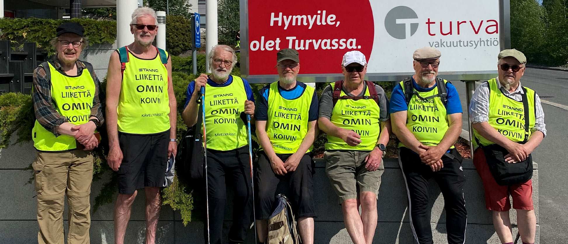 Seitsemän seniorimiestä hymyilevät auringonpaisteessa.
