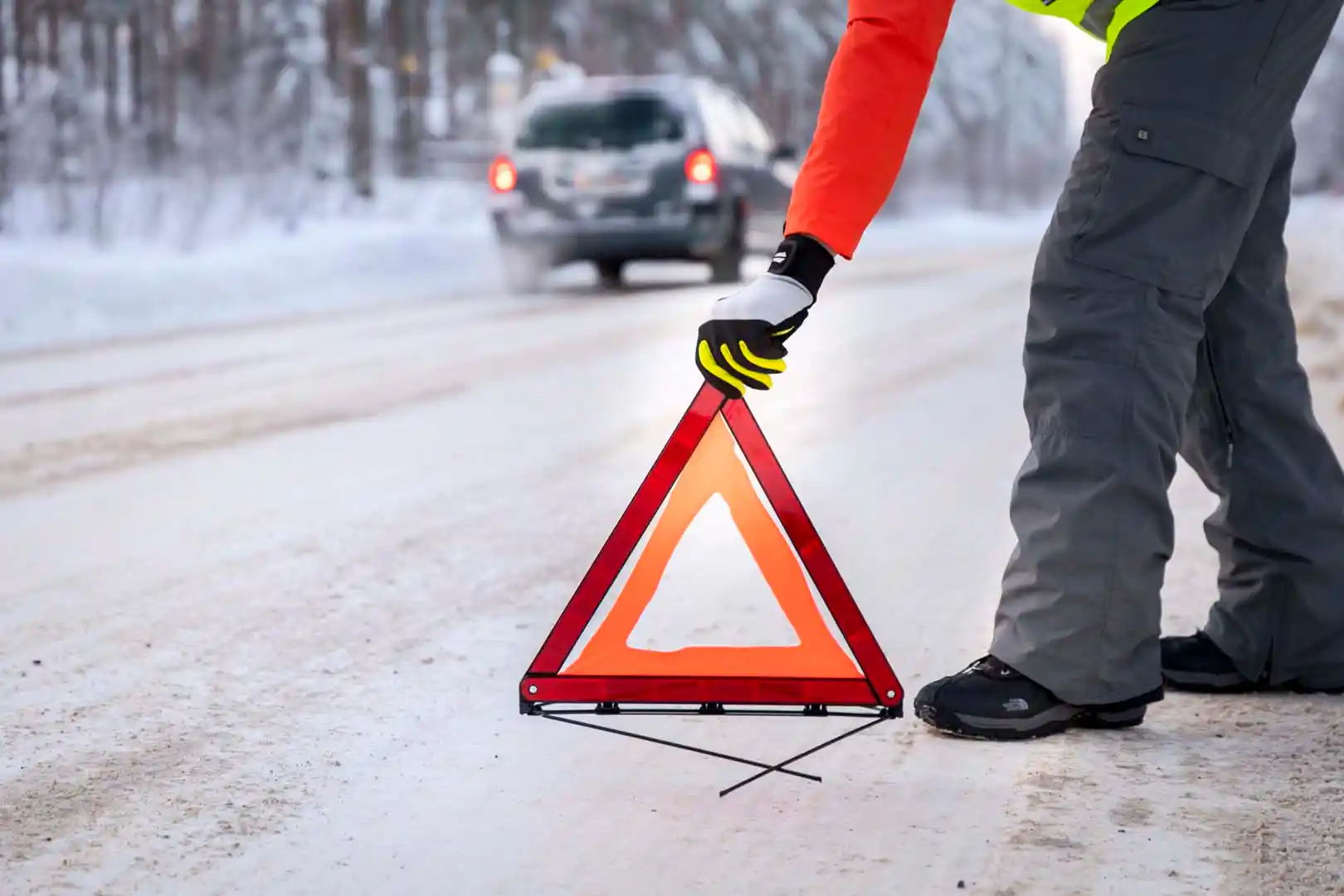 Harmaahousuinen, punapaitainen henkilö asettaa varoituskolmiota lumisen tien reunaan talvimaisemassa, taustalla näkyy ohiajavan harmaan henkilöauton perävalot.