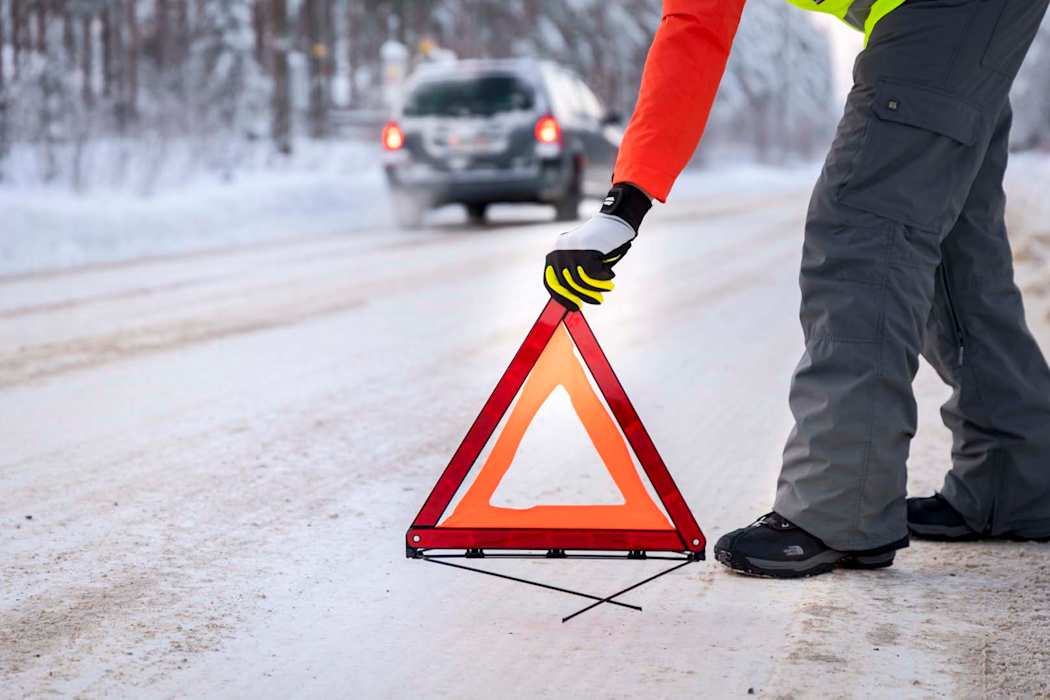 Harmaahousuinen, punapaitainen henkilö asettaa varoituskolmiota lumisen tien reunaan talvimaisemassa, taustalla näkyy ohiajavan harmaan henkilöauton perävalot.