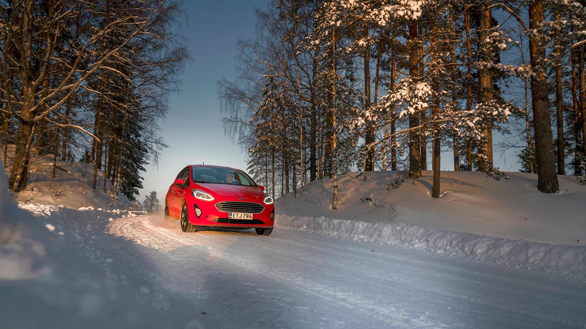 Punainen auto ajaa lumihankien keskellä hämärällä.