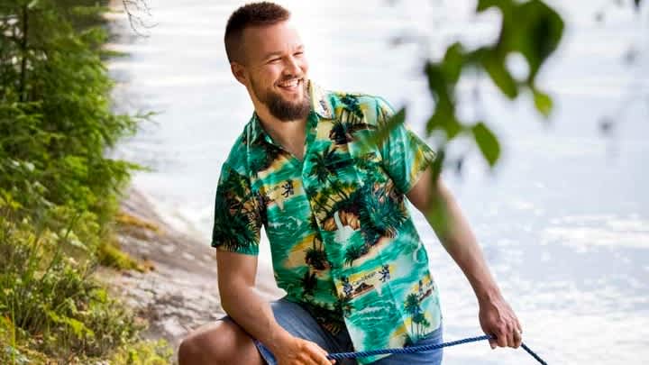 Kesävaatteisiin pukeutunut mies seisoo järven rannassa ja hymyilee.