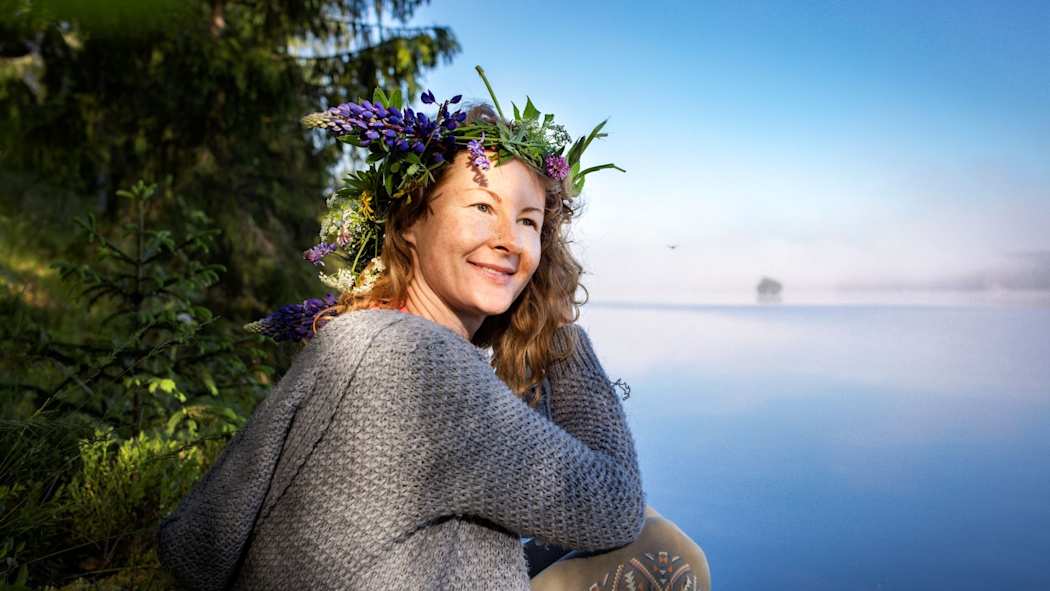 Nainen hymyilee kukkaseppele päässä järven rannassa.