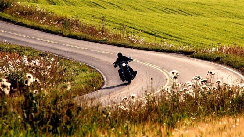 Ihminen ajaa moottoripyörällä aurinkoisena päivänä pellon keskellä mutkittelevalla maantiellä.